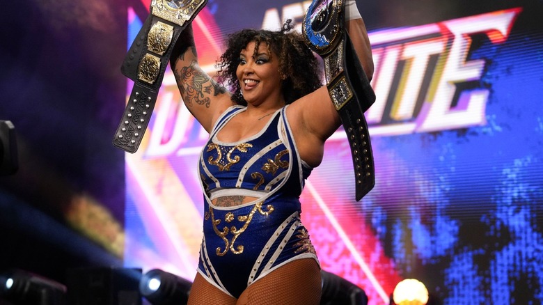 Willow Nightingale sticks her tongue out as she hoists up the AEW Women's World Tag Team Championship and the AEW TBS Championship.