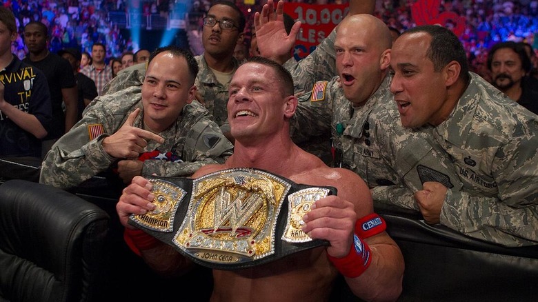 John Cena posing with the WWE Championship alongside soldiers at ringside during a show.