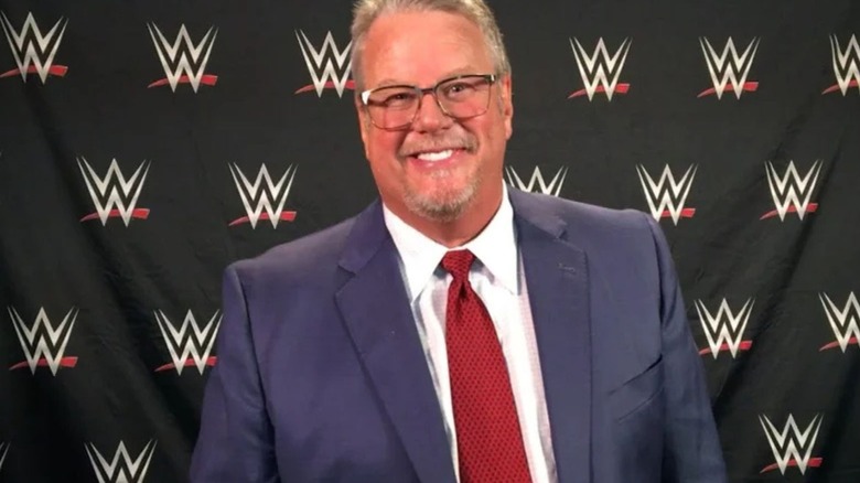Bruce Prichard smiles for a camera in front of a WWE-branded backdrop.